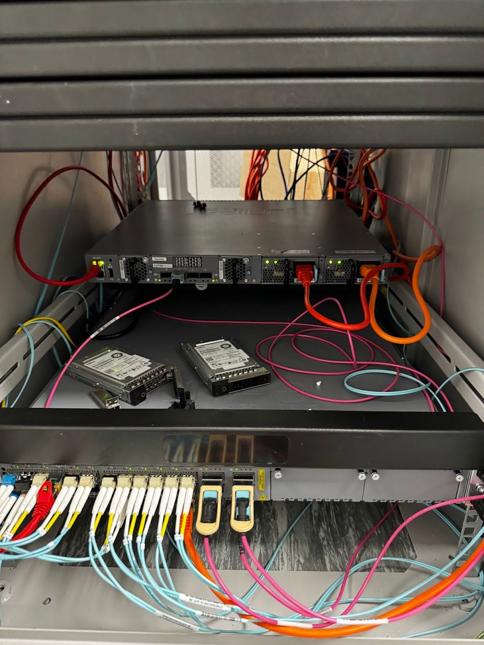 A 1U server pulled out on rails showing dual PSU, two hot-swap drives, and surrounding fibre patching